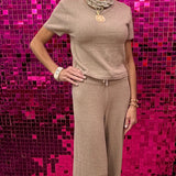 A woman modeling a beige knit short-sleeve top and matching wide-leg pants set, standing in front of a bright pink sequined wall.