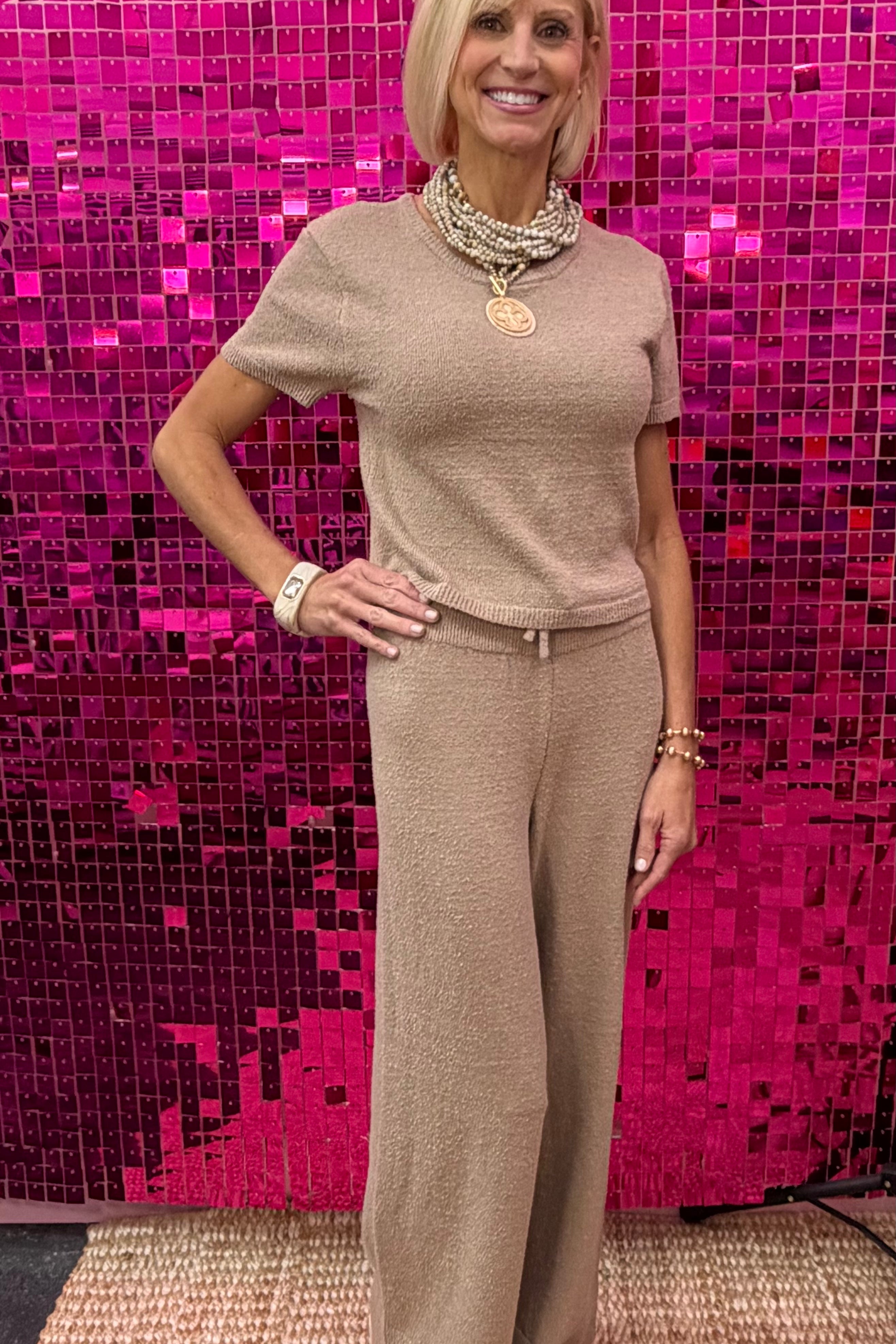 A woman modeling a beige knit short-sleeve top and matching wide-leg pants set, standing in front of a bright pink sequined wall.