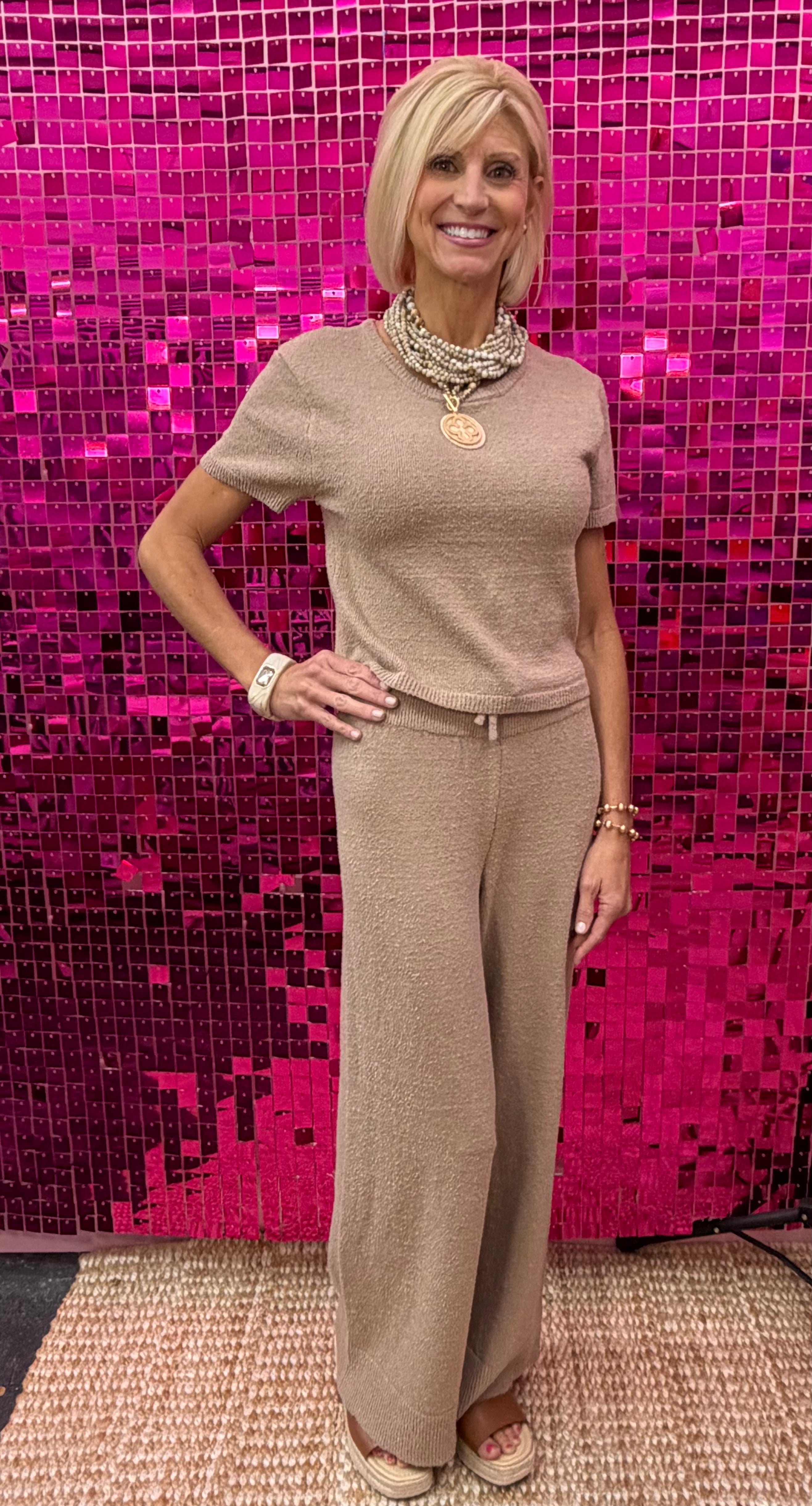 A woman modeling a beige knit short-sleeve top and matching wide-leg pants set, standing in front of a bright pink sequined wall.