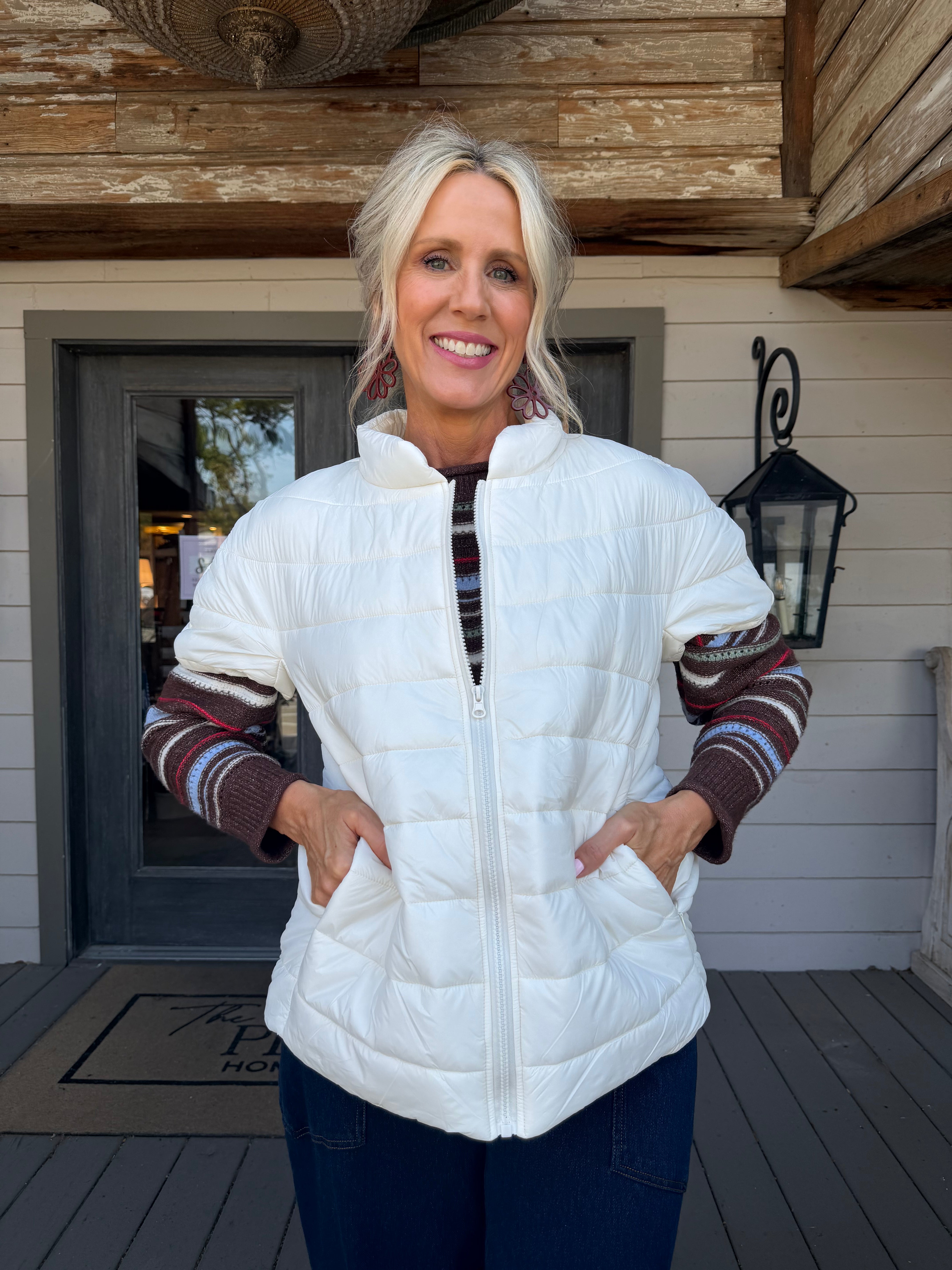 A smiling blonde woman stands on a porch wearing a white quilted zip-front puffer vest over a striped knit sweater and dark blue jeans.